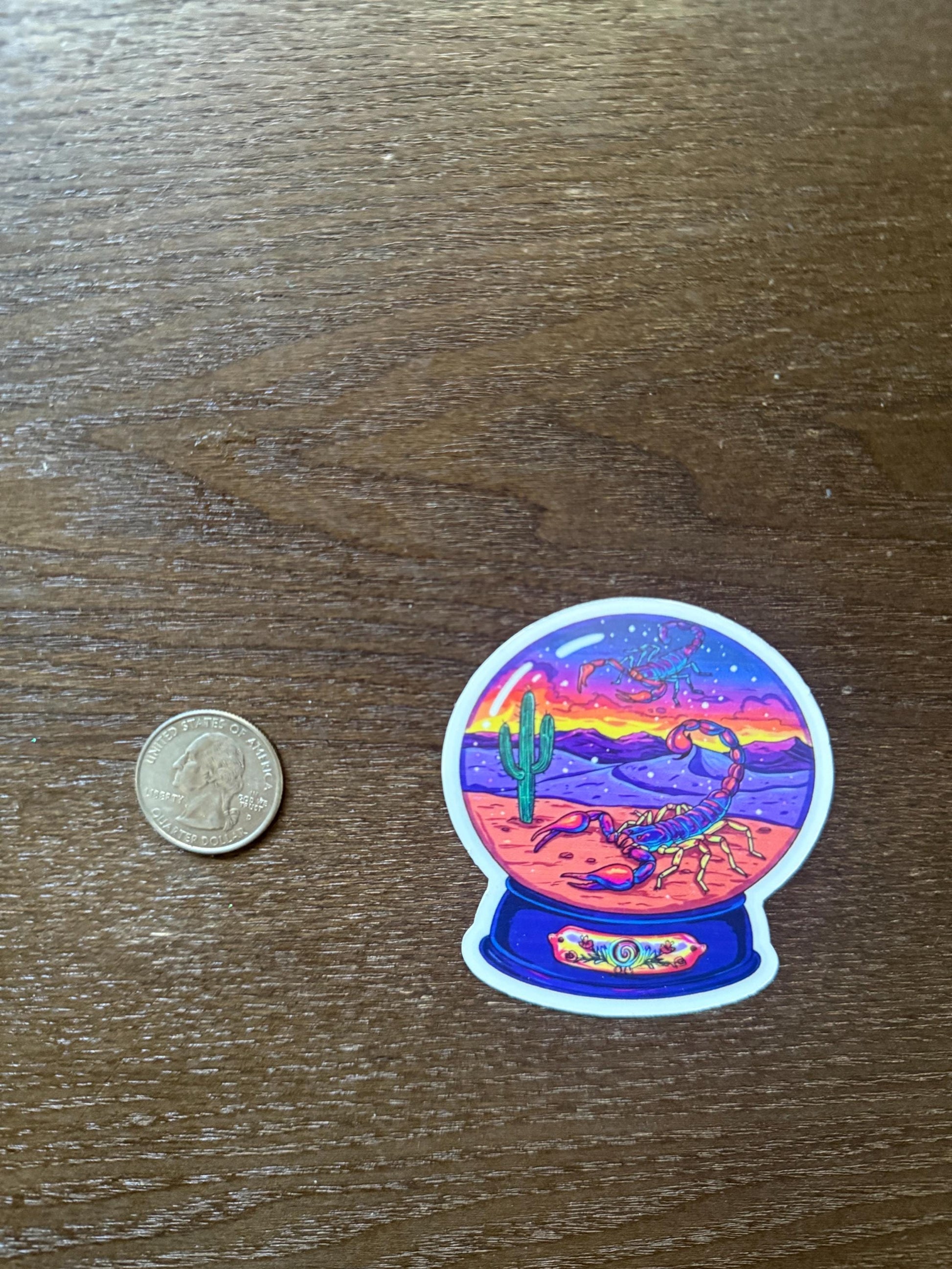 Sticker of a desert scene with a scorpion inside a crystal ball on a wooden surface, next to a coin for scale.