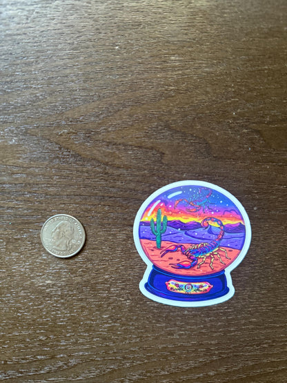 Sticker of a desert scene with a scorpion inside a crystal ball on a wooden surface, next to a coin for scale.