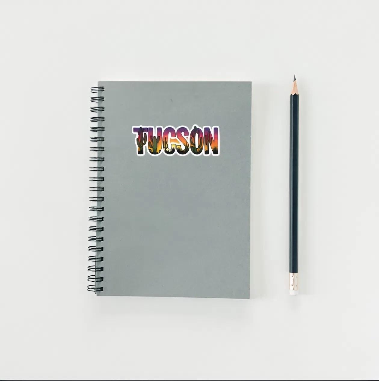 Gray notebook with 'TUCSON' in colorful letters and a pencil on a light gray background