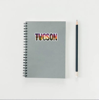 Gray notebook with 'TUCSON' in colorful letters and a pencil on a light gray background