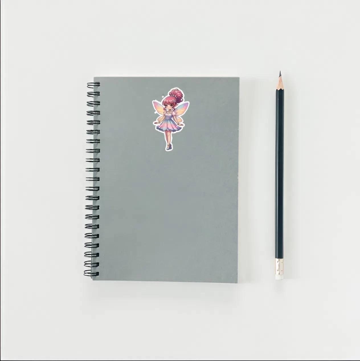 Gray spiral-bound notebook with a fairy sticker and a black pencil on a light gray background