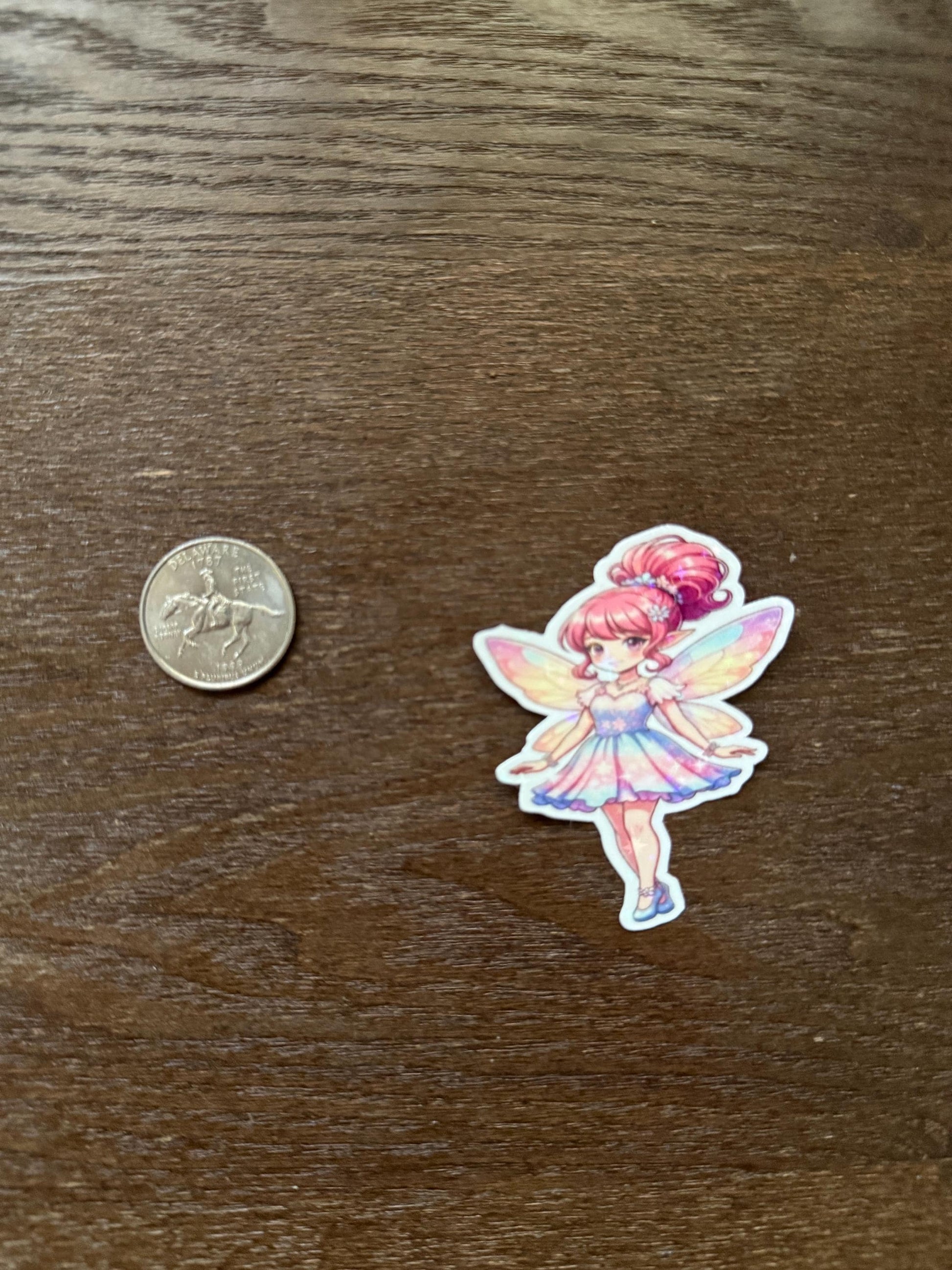 Sticker of a colorful fairy character next to a quarter on a wooden surface