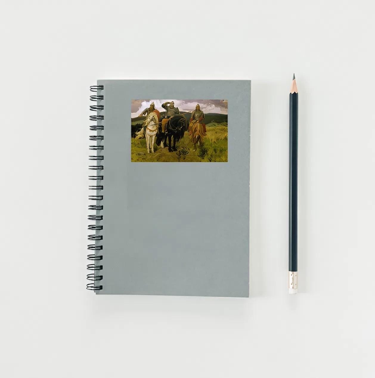 Notebook with a painting of people on horseback on its cover, next to a pencil on a white background