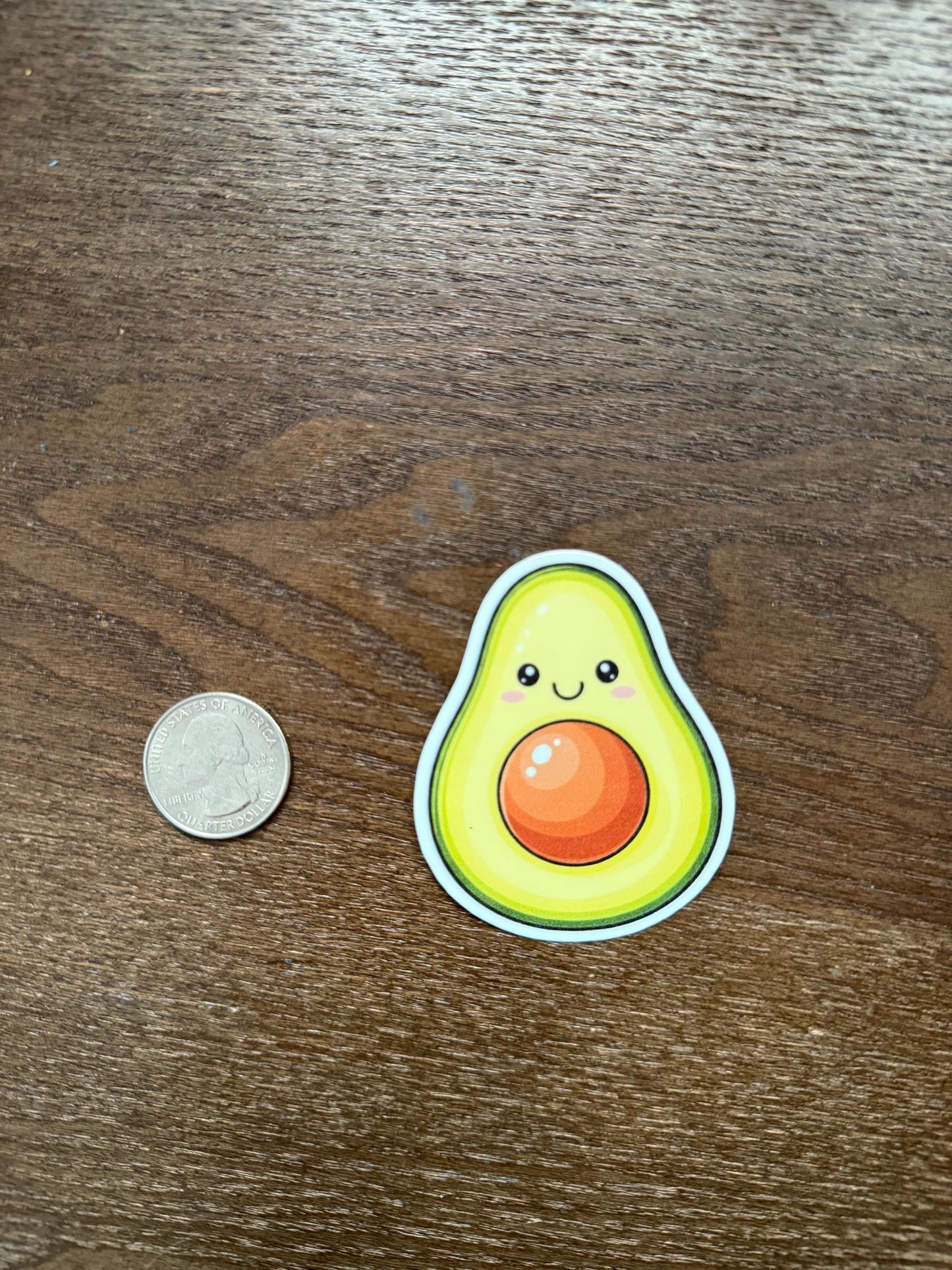 Sticker of a cute avocado sticker next to a coin on a wooden surface