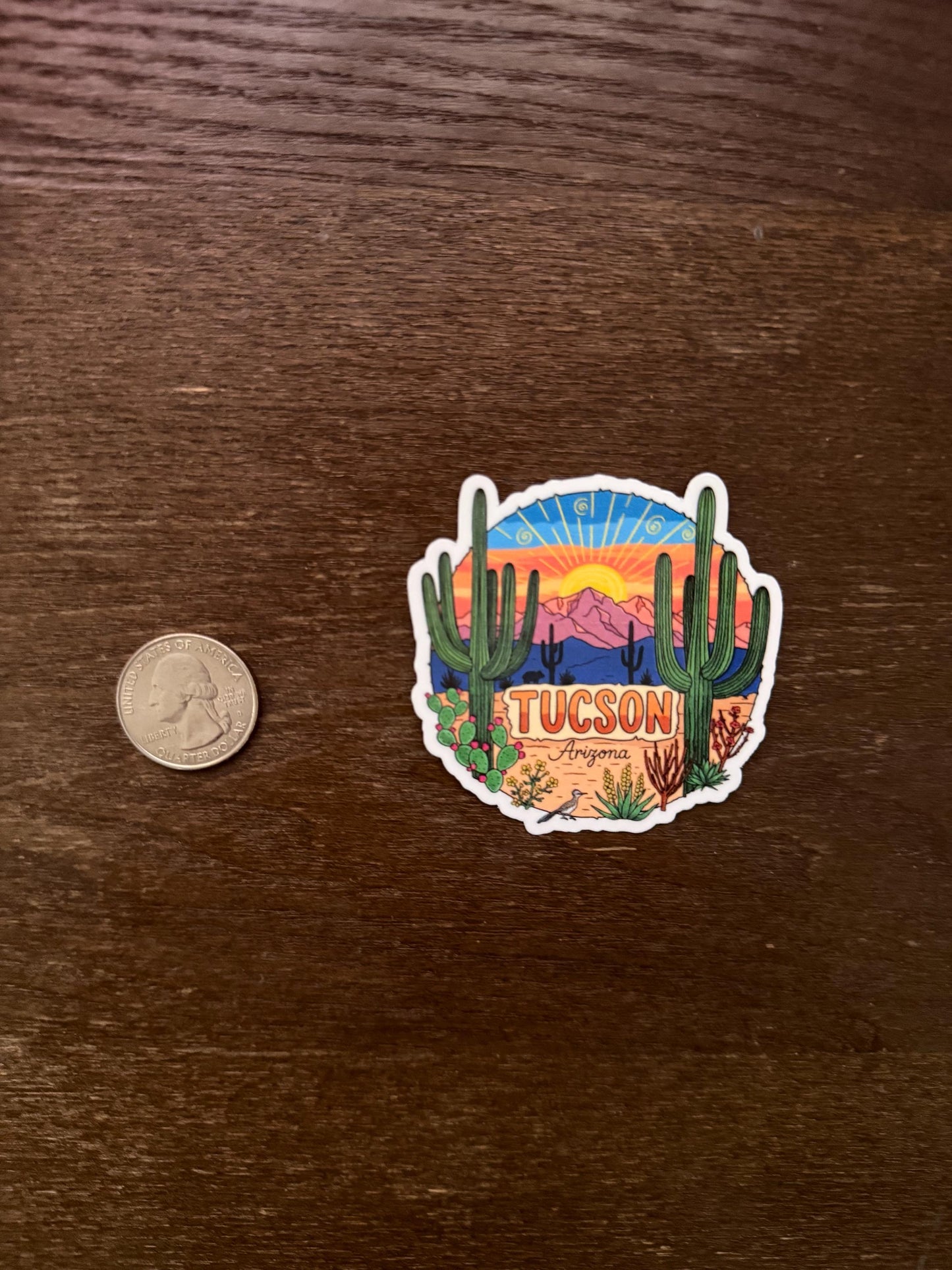 Sticker of Tucson, Arizona with a desert scene on a wooden surface next to a coin for scale.