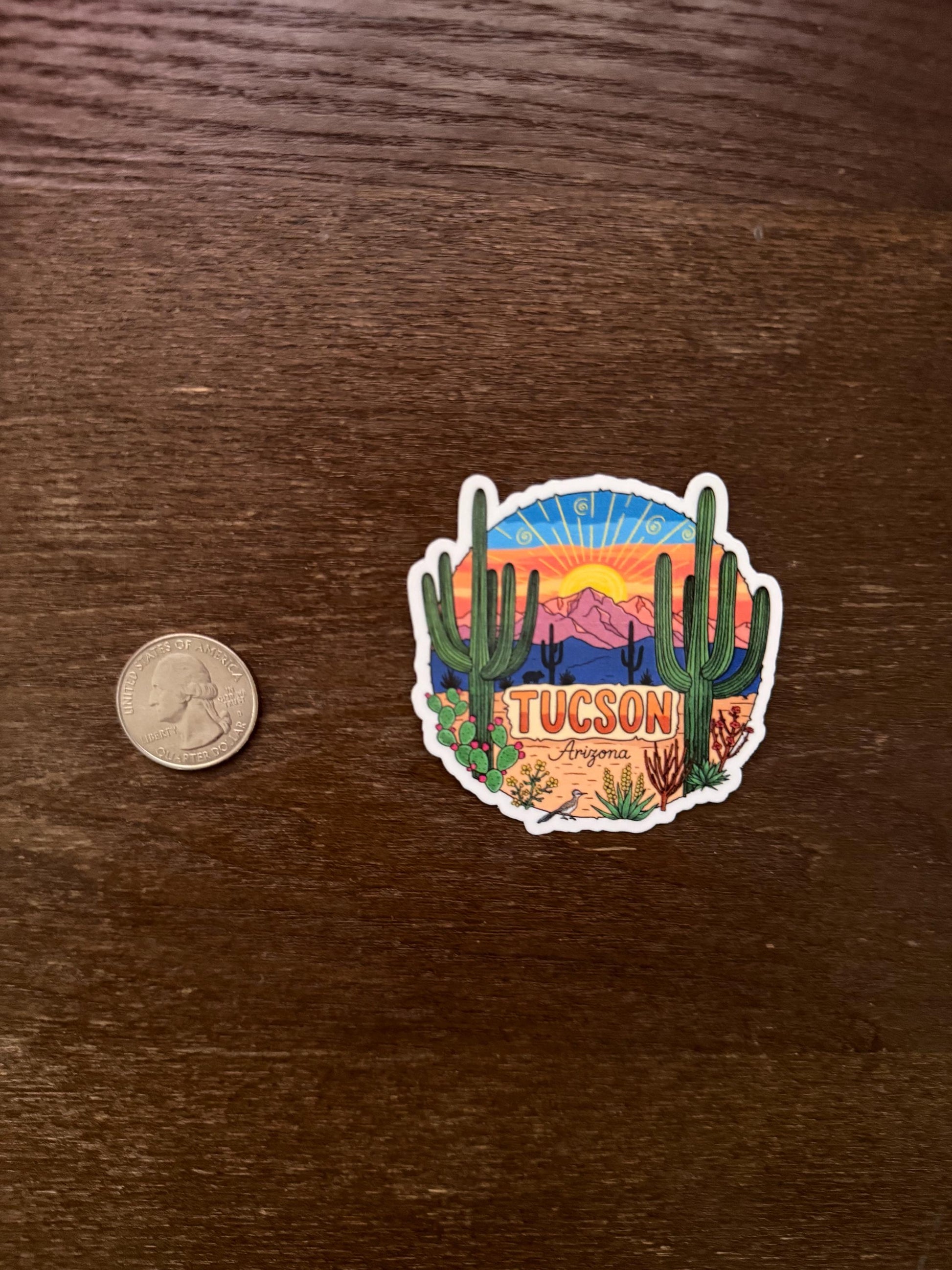 Sticker of Tucson, Arizona with a desert scene on a wooden surface next to a coin for scale.