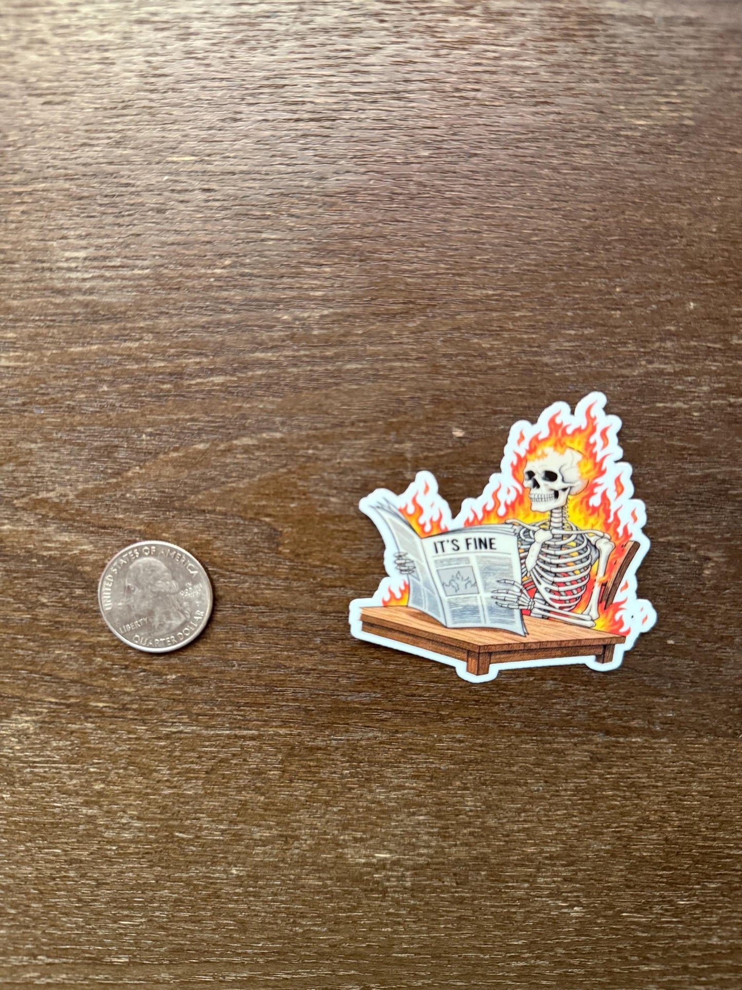 Sticker of a skeleton reading a newspaper with flames, next to a quarter for scale, on a wooden surface.