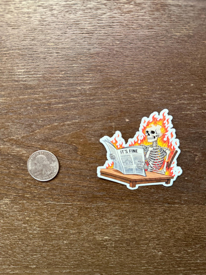 Sticker of a skeleton reading a newspaper with flames, next to a quarter for scale, on a wooden surface.