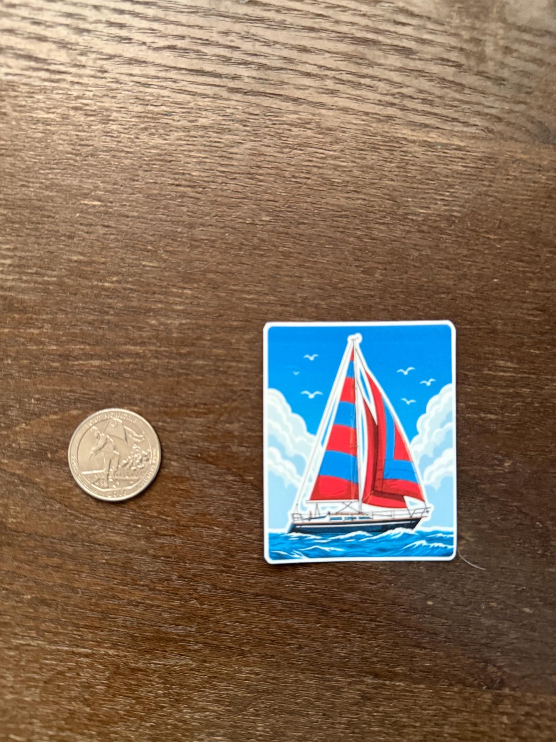 Sticker of a sailboat with red and blue sails on a wooden surface next to a coin for scale.