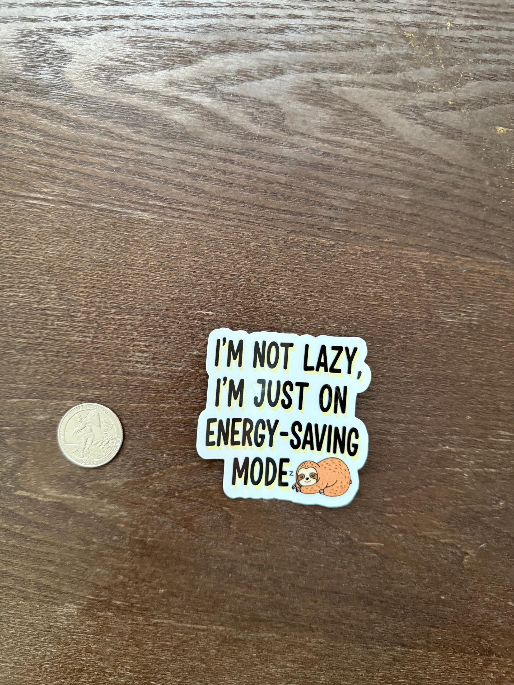 Sticker with text 'I'm not lazy, I'm just on energy-saving mode' on a wooden surface with a coin for scale.