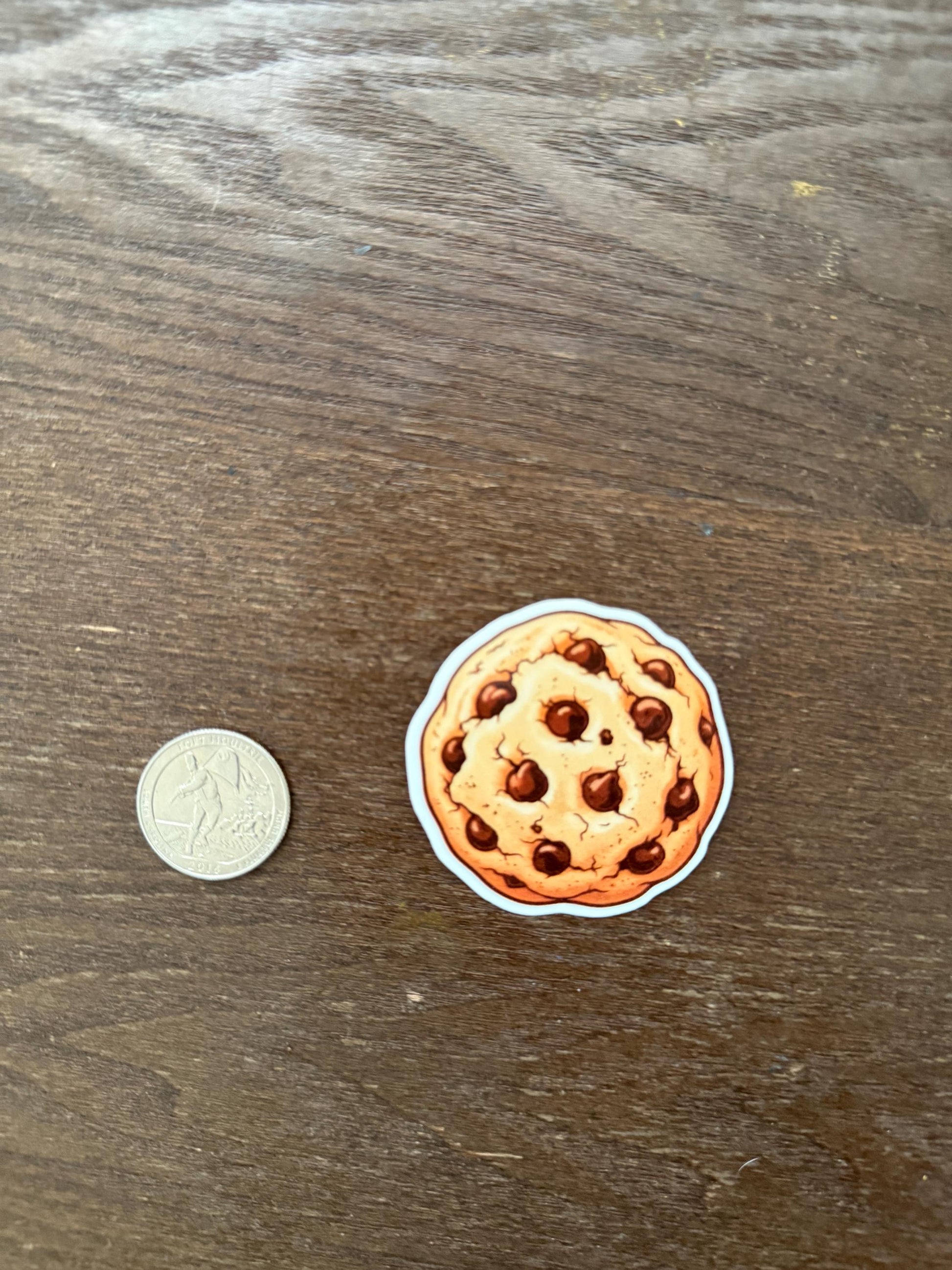 Sticker of a chocolate chip cookie next to a coin on a wooden surface