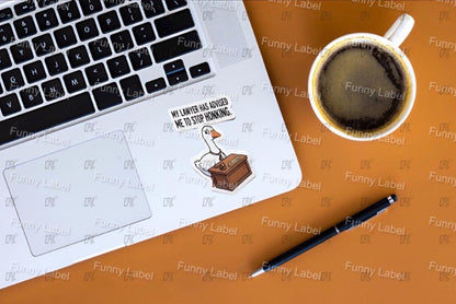 Laptop with a humorous sticker on a wooden surface next to a coffee cup and pen.