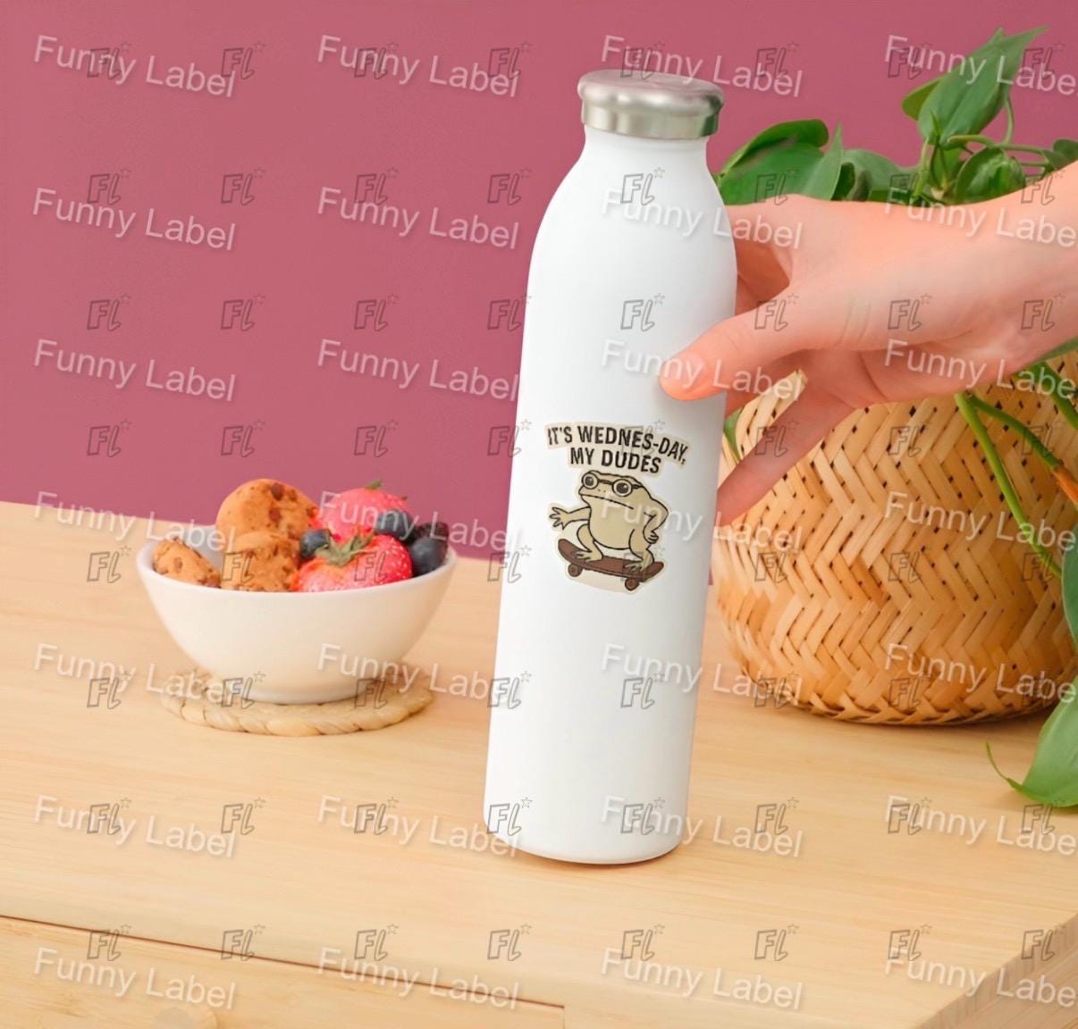 White water bottle with a design and text held by a hand on a wooden surface with a bowl of fruit and a plant.