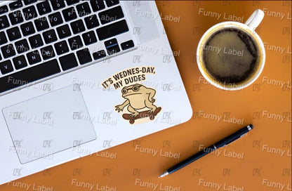Laptop with a 'It's Wednes-Day, My Dudes' sticker on an orange surface with a coffee cup and pen.