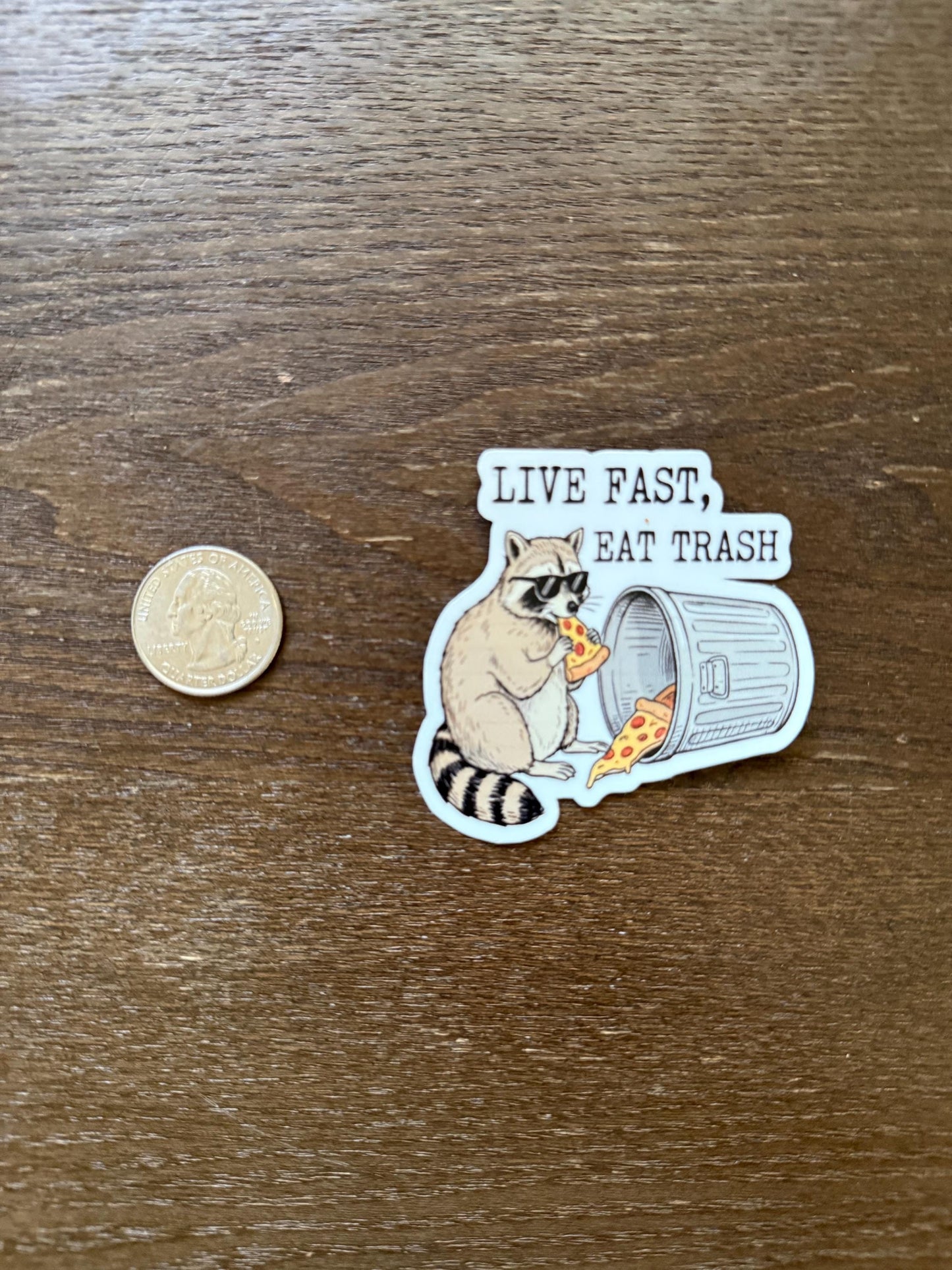 Sticker of a raccoon eating pizza with 'Live Fast, Eat Trash' text on a wooden surface.