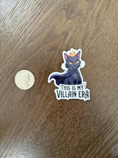 Sticker of a cat with a crown labeled 'This is my villain era' next to a coin on a wooden surface