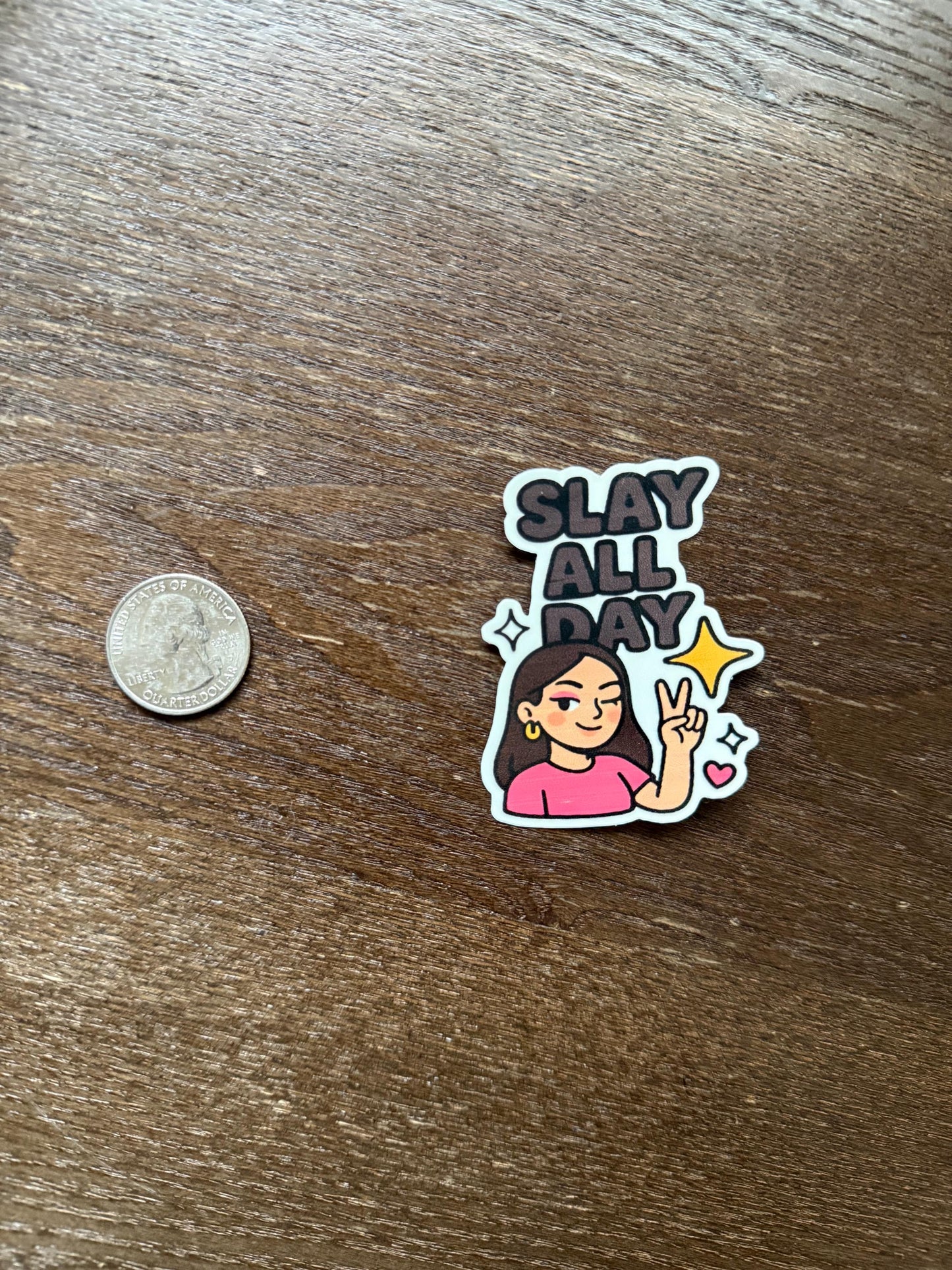 Sticker with 'Slay All Day' text and a person's illustration on a wooden surface with a coin for scale.