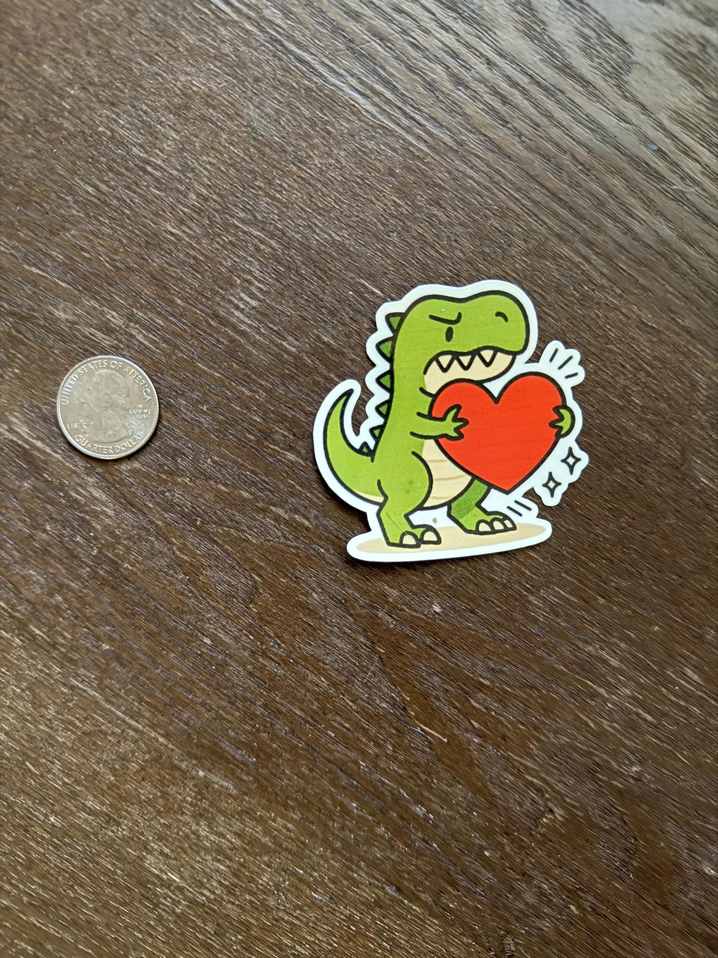 Sticker of a dinosaur holding a heart next to a coin on a wooden surface