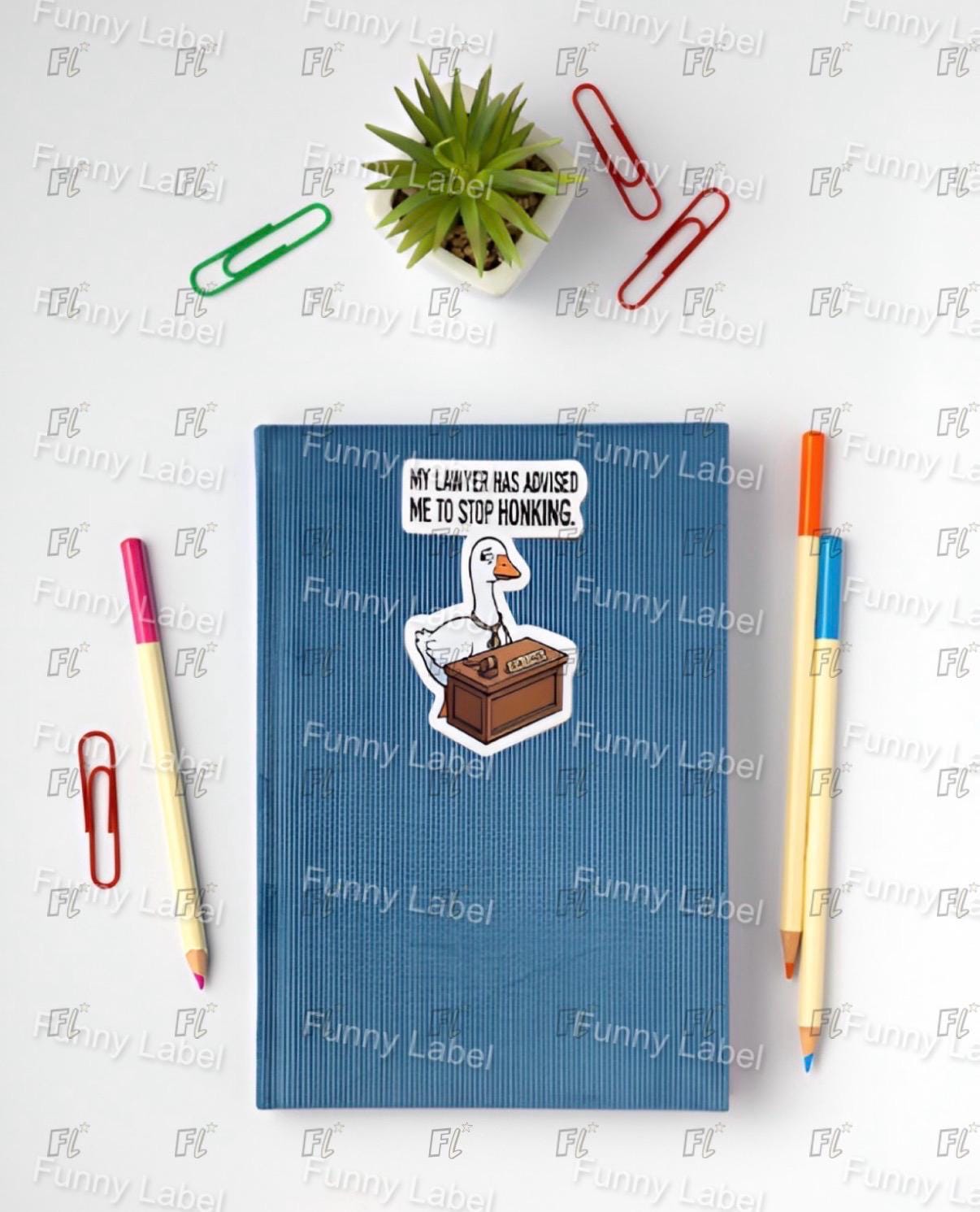 Blue notebook with a sticker of a goose and text, surrounded by office supplies on a white background.