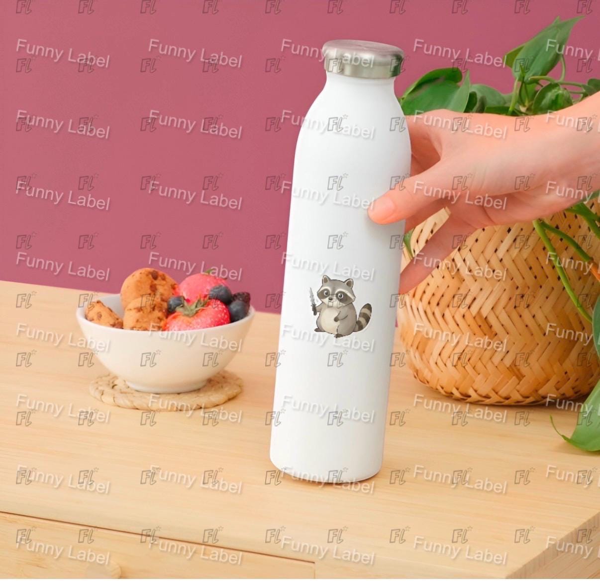 White water bottle with a raccoon design held by a hand on a wooden surface with a bowl of fruit and a basket.