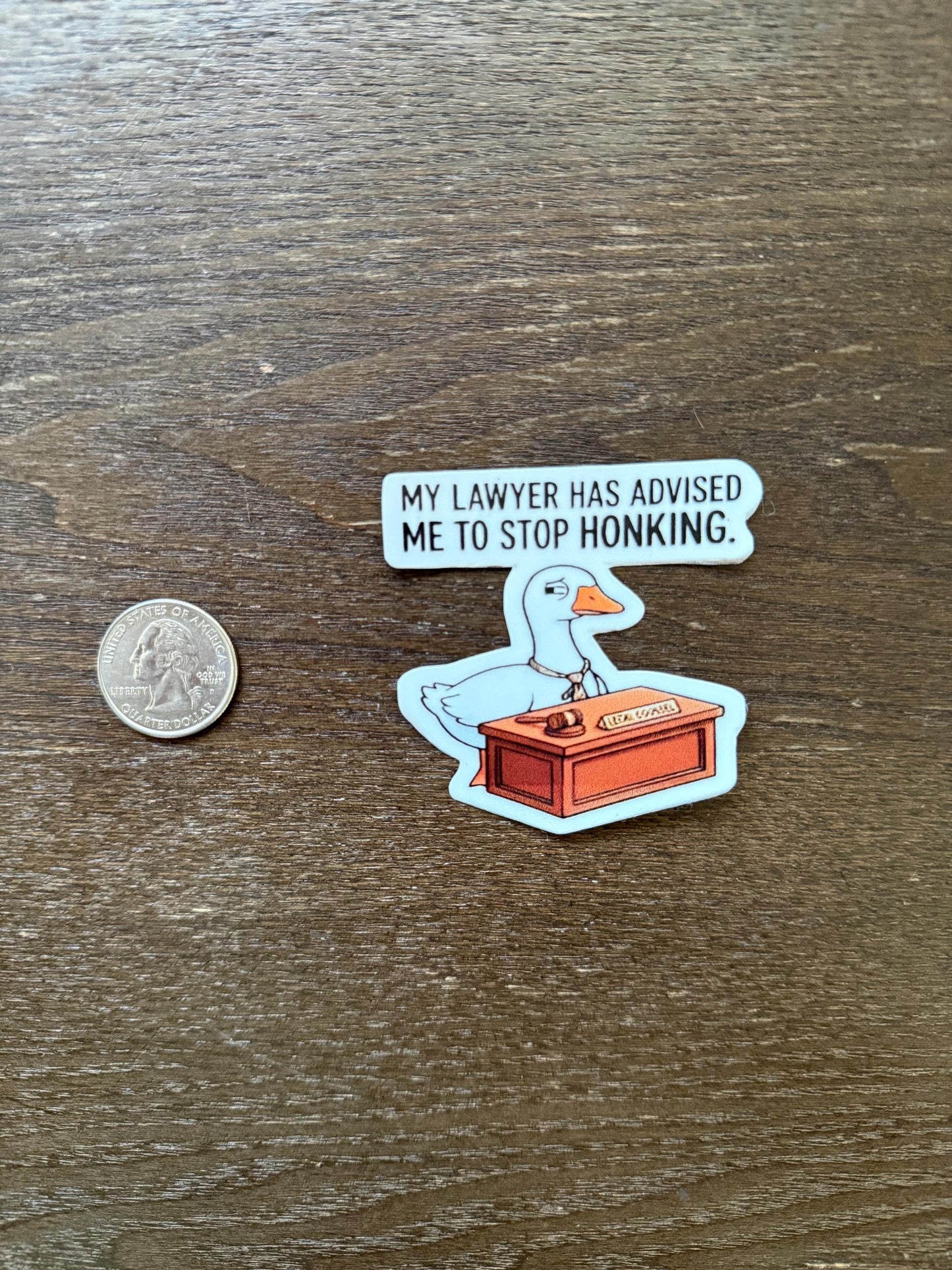 Sticker of a duck in a courtroom with a speech bubble and a coin for scale on a wooden surface