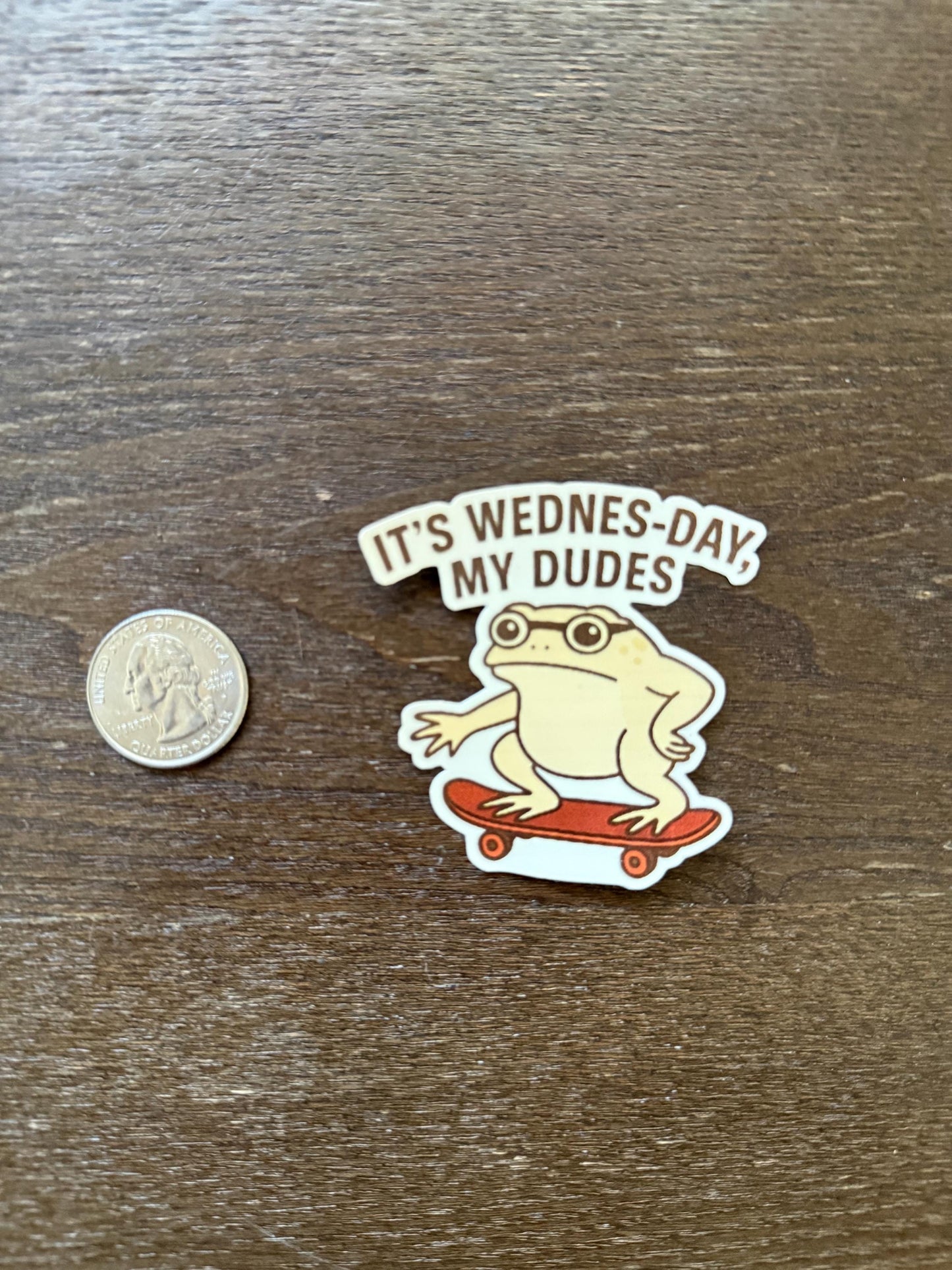 Sticker with a frog on a skateboard and text 'It's Wednes-Day My Dudes' next to a coin for scale on a wooden surface.