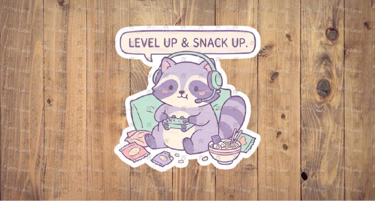 Sticker of a purple raccoon playing video games with text 'Level Up & Snack Up' on a wooden background