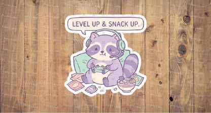 Sticker of a purple raccoon playing video games with text 'Level Up & Snack Up' on a wooden background