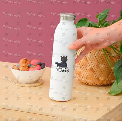 White water bottle with black cat design held by a hand on a wooden table with a pink background.